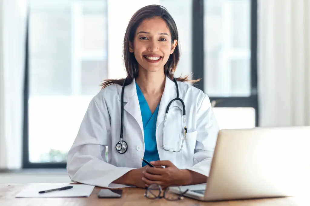 medica-consultorio-laptop-sonriente Médica en consultorio utilizando laptop, representando atención profesional y confianza en servicios de salud digitales.